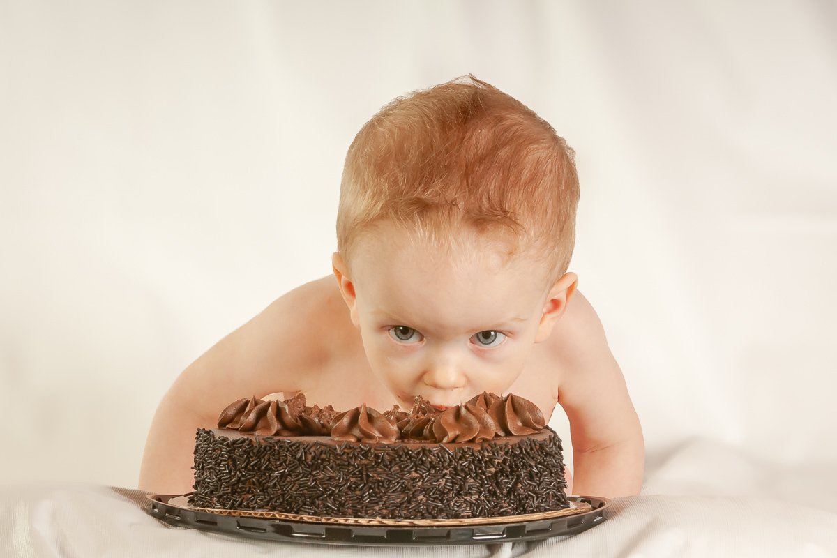 Baby leaning over a chocolate cake with an intensely curious expression, surrounded by a soft white backdrop.