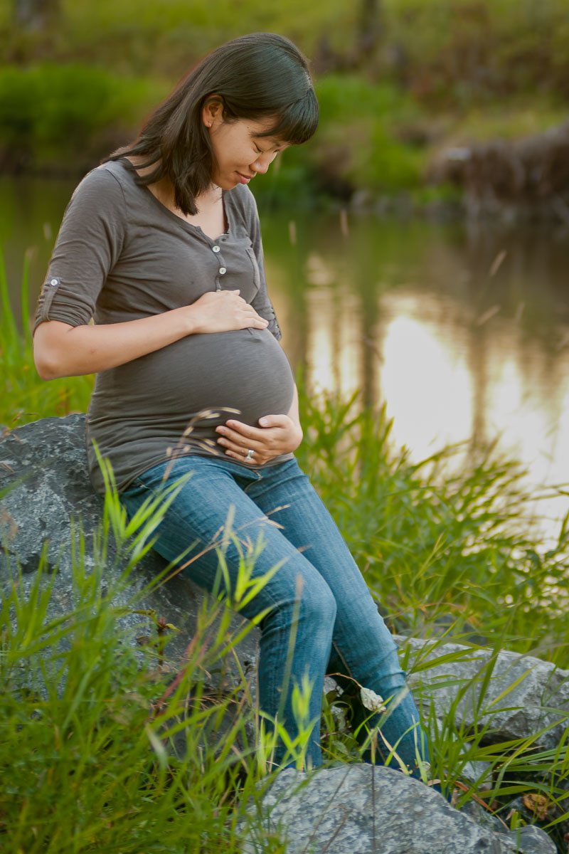 Expectant Mother sitting on rock next to water edge contemplating her baby bump