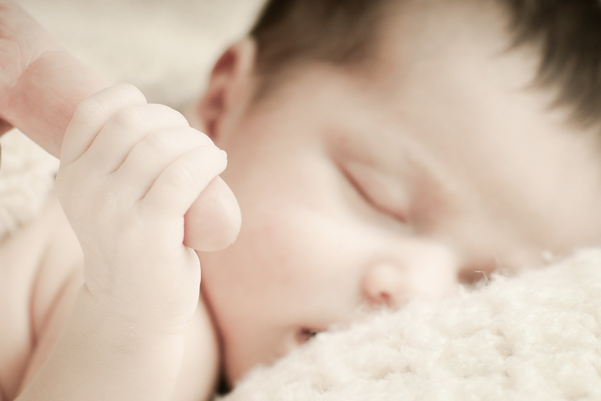 Sleeping newborn gently gripping an adult’s finger, with the baby’s tiny hand in sharp focus and the face softly blurred in the background.